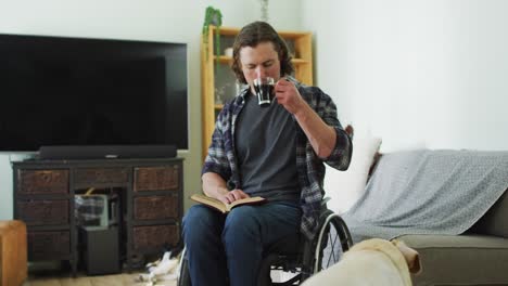 Relaxed-caucasian-disabled-man-in-wheelchair-reading-book-and-drinking-coffee-in-living-room