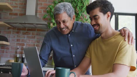 Smiling-biracial-man-with-arm-around-young-son-using-laptop-at-home
