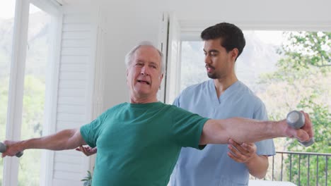 Young-male-physiotherapist-helping-retired-senior-man-exercising-with-dumbbells-at-nursing-home