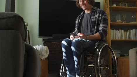 Glücklicher-Kaukasischer-Behinderter-Mann-Im-Rollstuhl,-Der-Sein-Smartphone-Im-Wohnzimmer-Benutzt