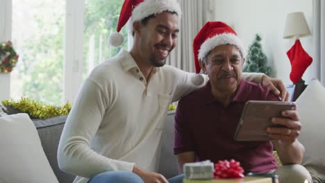 Cheerful-biracial-son-with-senior-father-in-santa-hats-making-video-call-on-tablet-pc-in-christmas