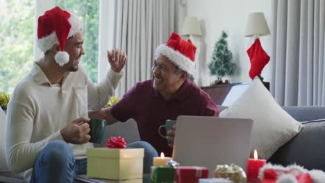 Happy-father-and-son-in-santa-hats-embracing-while-having-coffee-during-laptop-christmas-video-call