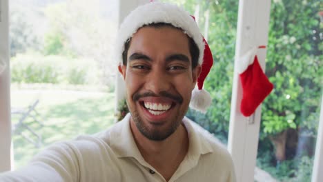 Portrait-of-cheerful-biracial-young-man-in-santa-hat-gesturing-during-christmas-video-call