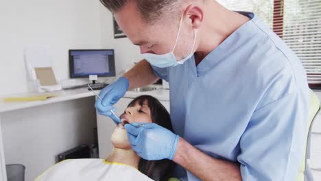 Caucasian-male-dentist-with-face-mask-examining-teeth-of-female-patient-at-modern-dental-clinic