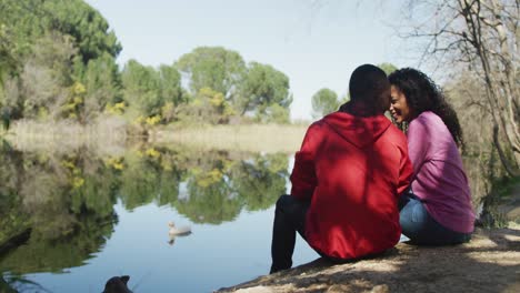 Smiling-diverse-couple-embracing-and-sitting-by-lake-in-countryside
