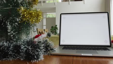 Laptop-with-copy-space-on-screen-on-table-with-santa-hats,-decorated-christmas-tree-and-presents