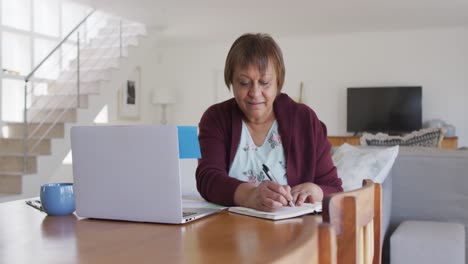 Lächelnde-Afroamerikanische-ältere-Frau,-Die-Am-Esstisch-Sitzt,-Einen-Laptop-Benutzt-Und-In-Ein-Notizbuch-Schreibt