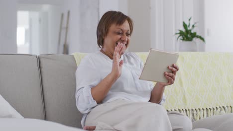 Happy-african-american-senior-woman-sitting-on-couch-making-video-call-using-tablet,-waving