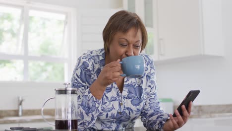 Feliz-Mujer-Mayor-Afroamericana-Disfrutando-Bebiendo-Una-Taza-De-Café-Y-Usando-Un-Teléfono-Inteligente