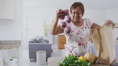 Glückliche-Afroamerikanische-Seniorin-In-Der-Küche,-Die-Lächelnd-Lebensmittel-Aus-Der-Einkaufstasche-Auspackt