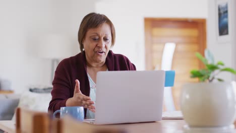 Feliz-Mujer-Mayor-Afroamericana-En-La-Mesa-Del-Comedor,-Haciendo-Videollamadas-Usando-Una-Computadora-Portátil-Y-Hablando