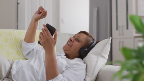 Happy-african-american-senior-woman-wearing-headphone-lying-on-couch-using-smartphone-and-smiling