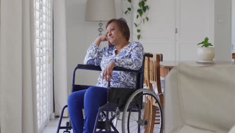 Happy-african-american-senior-woman-in-wheelchair-looking-out-of-window-at-home-smiling