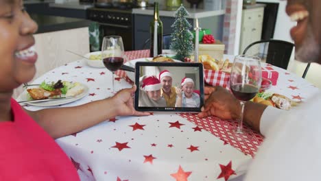 African-american-couple-with-wine-using-tablet-for-christmas-video-call-with-happy-family-on-screen