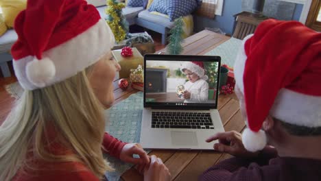 Smiling-caucasian-couple-with-santa-hats-using-laptop-for-christmas-video-call,-with-boy-on-screen