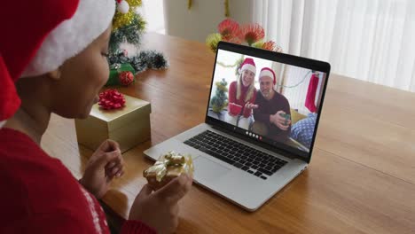 Afroamerikanische-Frau-Mit-Weihnachtsmütze-Nutzt-Laptop-Für-Weihnachtsvideoanruf-Mit-Paar-Auf-Dem-Bildschirm