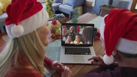 Lächelndes-Kaukasisches-Paar-Mit-Weihnachtsmützen,-Das-Einen-Laptop-Für-Einen-Weihnachtsvideoanruf-Mit-Der-Familie-Auf-Dem-Bildschirm-Benutzt