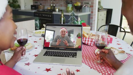 Smiling-african-american-couple-with-wine-using-laptop-for-christmas-video-call-with-man-on-screen