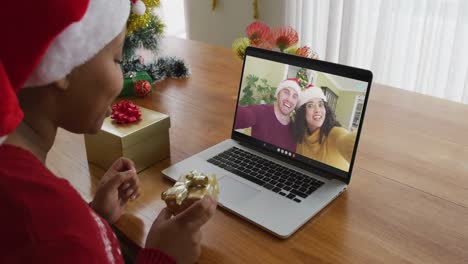 African-american-woman-with-santa-hat-using-laptop-for-christmas-video-call,-with-couple-on-screen