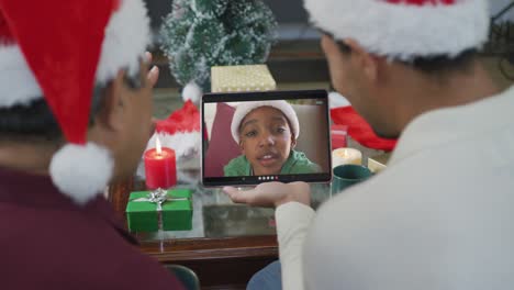 Birazialer-Vater-Und-Sohn-Mit-Weihnachtsmützen,-Die-Ein-Tablet-Für-Einen-Weihnachtsvideoanruf-Mit-Einem-Jungen-Auf-Dem-Bildschirm-Nutzen
