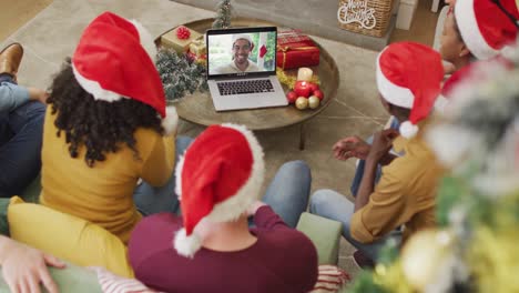 Diverse-family-with-santa-hats-using-laptop-for-christmas-video-call-with-smiling-man-on-screen
