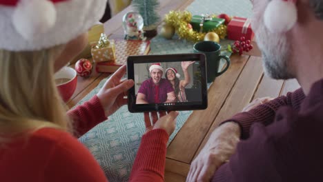 Lächelndes-Kaukasisches-Paar-Mit-Weihnachtsmützen,-Das-Ein-Tablet-Für-Einen-Weihnachtsvideoanruf-Mit-Einem-Paar-Auf-Dem-Bildschirm-Verwendet