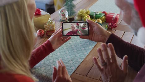 Kaukasisches-Paar-Mit-Weihnachtsmützen-Nutzt-Smartphone-Für-Weihnachtsvideoanruf-Mit-Mann-Auf-Dem-Bildschirm