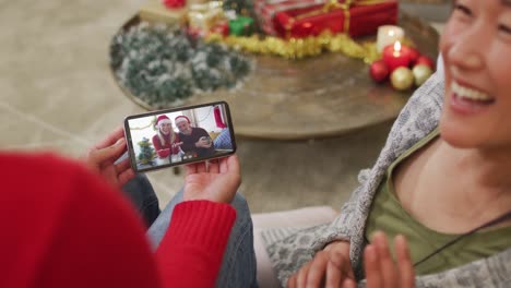 Smiling-asian-couple-using-smartphone-for-christmas-video-call-with-couple-with-santa-hats-on-screen