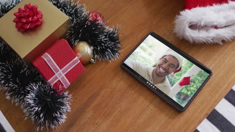 Smiling-biracial-man-wearing-santa-hat-on-christmas-video-call-on-tablet
