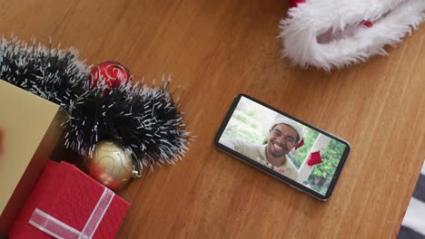 Smiling-biracial-man-wearing-santa-hat-on-christmas-video-call-on-smartphone