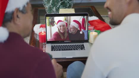 Lächelnder-Gemischtrassiger-Vater-Mit-Sohn,-Der-Einen-Laptop-Für-Einen-Weihnachtsvideoanruf-Mit-Einem-Paar-Auf-Dem-Bildschirm-Benutzt