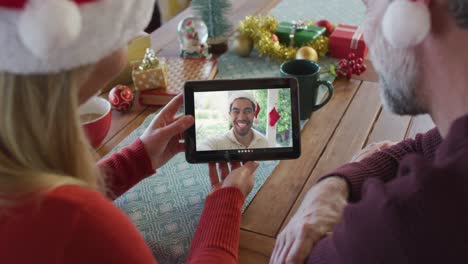 Lächelndes-Kaukasisches-Paar-Mit-Weihnachtsmützen,-Das-Ein-Tablet-Für-Einen-Weihnachtsvideoanruf-Mit-Einem-Mann-Auf-Dem-Bildschirm-Nutzt