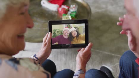 Smiling-senior-caucasian-couple-using-tablet-for-christmas-video-call-with-couple-on-screen