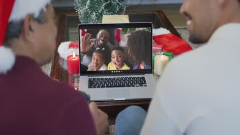 Biracial-father-and-son-with-santa-hats-using-laptop-for-christmas-video-call-with-family-on-screen