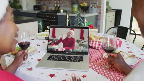 African-american-couple-with-wine-using-laptop-for-christmas-video-call-with-happy-man-on-screen