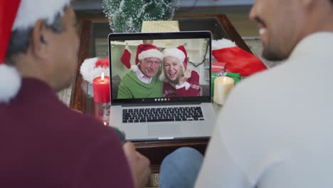 Smiling-biracial-father-with-son-using-laptop-for-christmas-video-call-with-senior-couple-on-screen