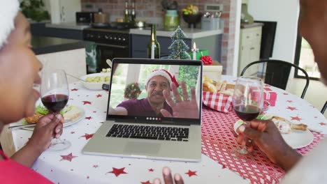 African-american-couple-with-wine-using-laptop-for-christmas-video-call-with-man-on-screen