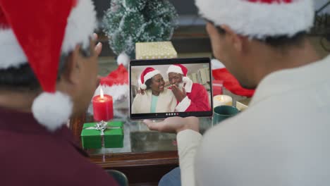 Biracial-father-and-son-with-santa-hats-using-tablet-for-christmas-video-call-with-family-on-screen