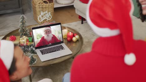 Vielfältige-Familie-Mit-Weihnachtsmützen-Nutzt-Laptop-Für-Weihnachtsvideoanruf-Mit-Glücklichem-Weihnachtsmann-Auf-Dem-Bildschirm