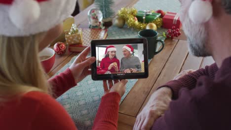 Caucasian-couple-with-santa-hats-using-tablet-for-christmas-video-call-with-smiling-family-on-screen