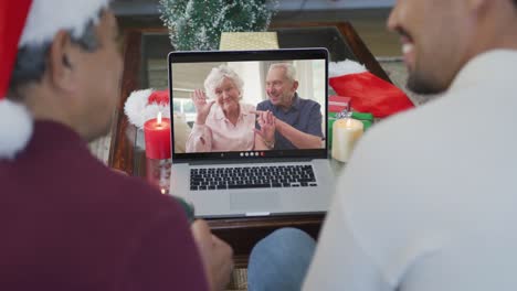 Lächelnder,-Gemischtrassiger-Vater-Und-Sohn,-Die-Einen-Laptop-Für-Einen-Weihnachtsvideoanruf-Mit-Einem-älteren-Paar-Auf-Dem-Bildschirm-Nutzen