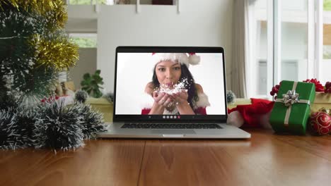 Smiling-caucasian-woman-wearing-santa-hat-and-blowing-snow-on-christmas-video-call-on-laptop