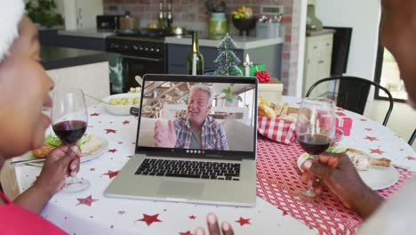 African-american-couple-with-wine-using-laptop-for-christmas-video-call-with-happy-man-on-screen