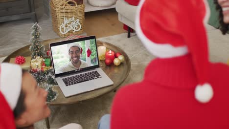 Diverse-family-with-santa-hats-using-laptop-for-christmas-video-call-with-happy-man-on-screen