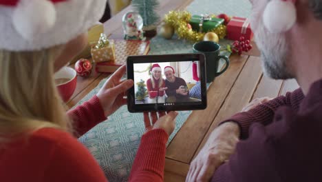 Smiling-caucasian-couple-with-santa-hats-using-tablet-for-christmas-video-call-with-couple-on-screen