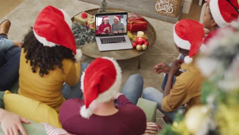 Diverse-family-with-santa-hats-using-laptop-for-christmas-video-call-with-smiling-couple-on-screen