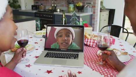 African-american-couple-with-wine-using-laptop-for-christmas-video-call-with-boy-on-screen