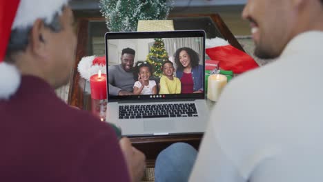 Smiling-biracial-father-with-son-using-laptop-for-christmas-video-call-with-family-on-screen