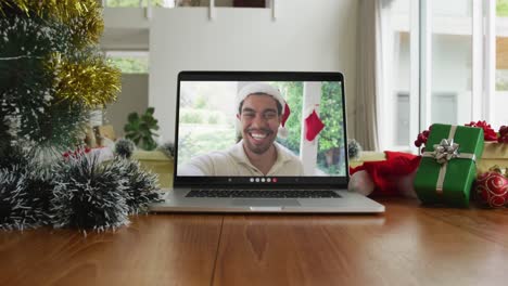 Smiling-biracial-man-wearing-santa-hat-on-christmas-video-call-on-laptop