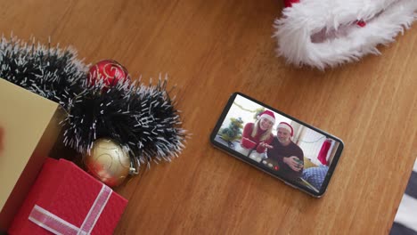 Smiling-caucasian-couple-wearing-santa-hats-on-christmas-video-call-on-smartphone